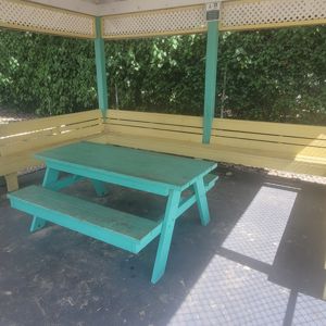 A shaded seating area with a teal table and benches under a structure surrounded by greenery.