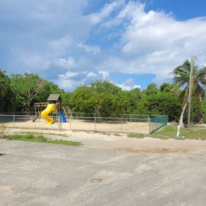 A playground with a yellow slide surrounded by greenery, showing clear blue sky and scattered clouds.