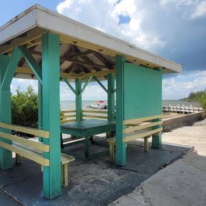 A gazebo by the waterfront, surrounded by green foliage and a clear sky.