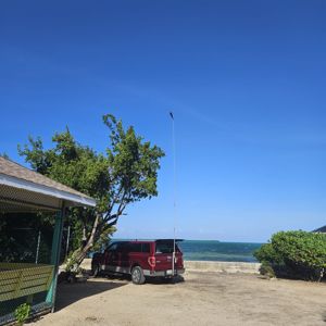 A view of a vehicle parked near the water with antennas set up, surrounded by trees and a clear blue sky.