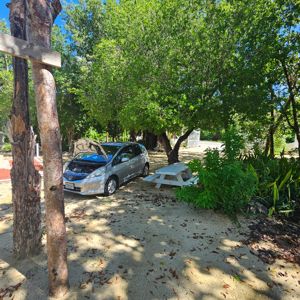 A parked silver car with its hood open under a shaded area with trees, along with a white picnic table nearby.