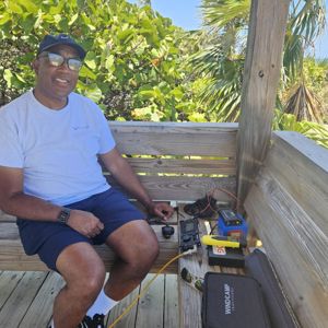 Operator seated at wooden platform with equipment, surrounded by greenery and a clear blue sky.