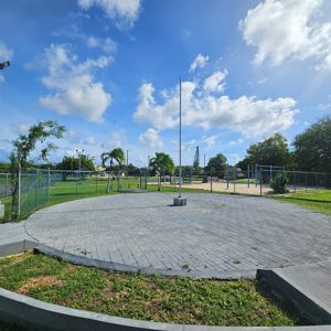 Open area in a park with pavement, palm trees, and a playground visible in the background under a blue sky with clouds.
