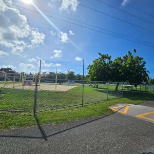 A sunny park area with a fenced playground, picnic tables, and trees.