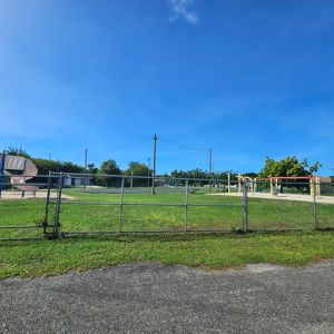 A wide view of a park area with a playground and basketball court, enclosed by a chain-link fence and under a clear blue sky.