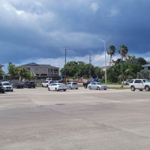 A parking area with several parked cars, surrounded by residential buildings and palm trees under partly cloudy skies.