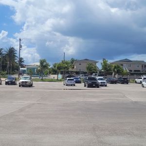 A parking area surrounded by palm trees and residential buildings under a partly cloudy sky.