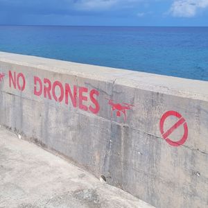A concrete barrier with a 'No Drones' sign painted in red, overlooking a blue ocean.