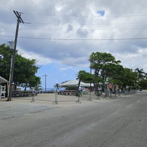 A view of a road lined with trees and temporary fencing with some structures in the background under a cloudy sky.