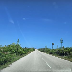 A scenic view of a winding road leading towards a bright blue sky, surrounded by lush greenery and trees.