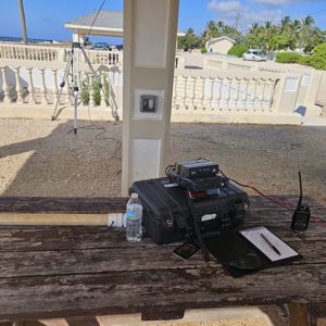 A radio setup on a wooden table under a shelter, with equipment visible, including a radio, notebook, and a water bottle, with a view of trees and a beach in the background.