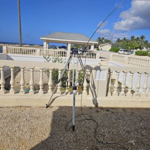 A tripod with an antenna is set up in a residential area, surrounded by a low wall and vegetation under a clear blue sky.