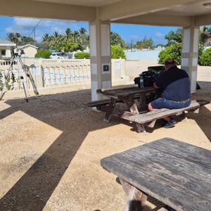 A radio operator seated at a wooden picnic table under a shelter, with equipment set up nearby.