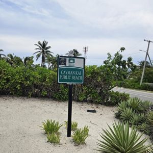 Sign for Cayman Kai Public Beach with surrounding greenery and sandy area.