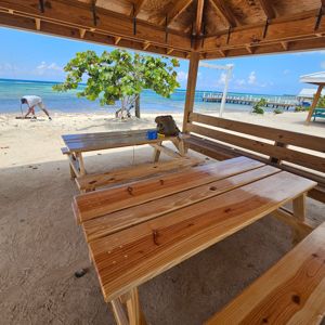 A shaded area with wooden picnic tables near the beach, showing a tree and a person in the background.