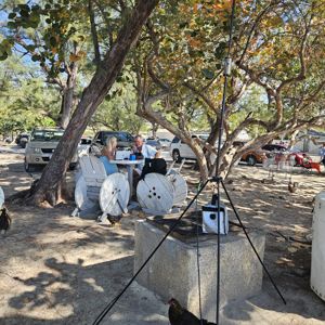 Operators set up at a shaded outdoor location with trees and a tripod antenna, surrounded by vehicles and wildlife.