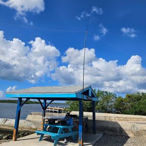A radio operator sitting at a blue picnic table under a shelter by the water, with an antenna setup.