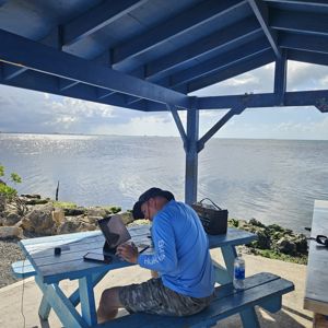 A radio operator working at a blue picnic table under a shaded pavilion by the waterfront.