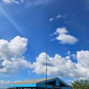 A clear blue sky with white clouds and a vertical antenna attached to a blue-roofed structure.
