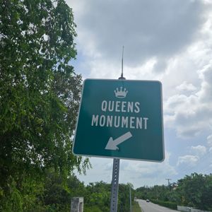 A sign indicating the direction to Queens Monument with lush greenery in the background.